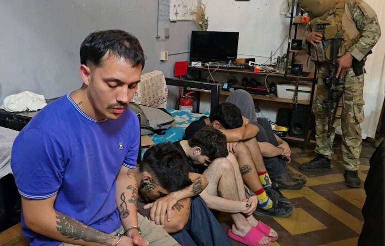 Cinco personas en ambiente interior, cuatro encadenadas y mirando al suelo con tensión, mientras un agente de seguridad en camiseta camuflada sostiene un arma.