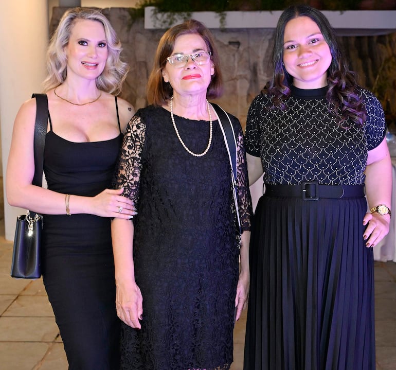 Tres mujeres sonríen en evento elegante: una en vestido negro ajustado, otra en encaje y collar de perlas, y la última en blusa brillante.
