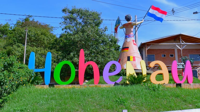 Hombre en vestimenta paraguaya sostiene la bandera de Paraguay frente a un colorido letrero que dice 'Hohenau', sobre césped verde.