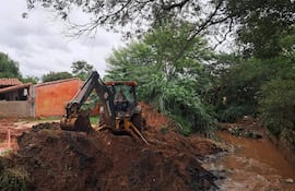 Hombre en retroexcavadora con camisa de cuadros trabaja en terreno de barro. Fondo con casas y vegetación.