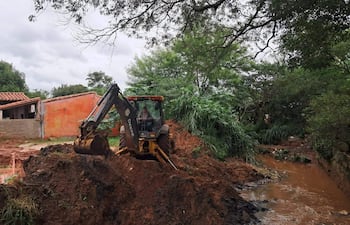 Hombre en retroexcavadora con camisa de cuadros trabaja en terreno de barro. Fondo con casas y vegetación.