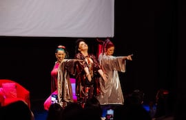 Tres mujeres en trajes coloridos sobre el escenario, una de ellas con cabello verde, otra con cabello rojo, actuando con confianza.