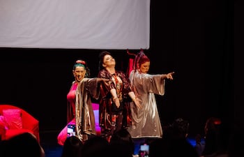Tres mujeres en trajes coloridos sobre el escenario, una de ellas con cabello verde, otra con cabello rojo, actuando con confianza.