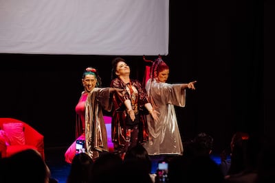 Tres mujeres en trajes coloridos sobre el escenario, una de ellas con cabello verde, otra con cabello rojo, actuando con confianza.