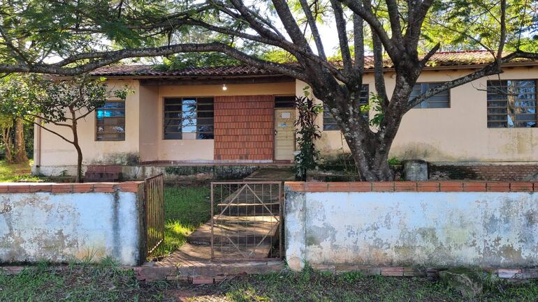 Casa beige con techo de tejas en un entorno natural, rodeada de césped y un árbol grande.