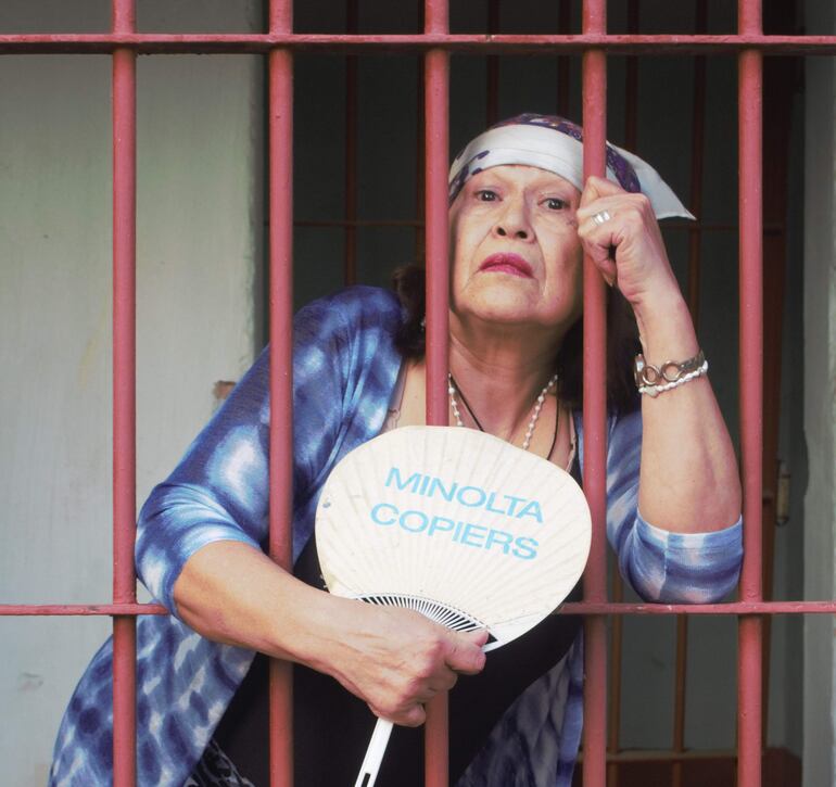 Mujer en atuendo azul con banda blanca sosteniendo un abanico con logo, expresando descontento.