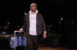 Antonio Sanabria, vestido con abrigo gris y sudadera blanca, camina en un ambiente nocturno, sosteniendo una maleta y un maletín.