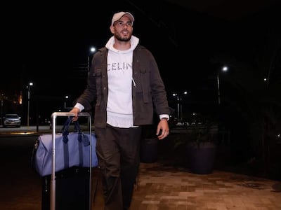 Antonio Sanabria, vestido con abrigo gris y sudadera blanca, camina en un ambiente nocturno, sosteniendo una maleta y un maletín.