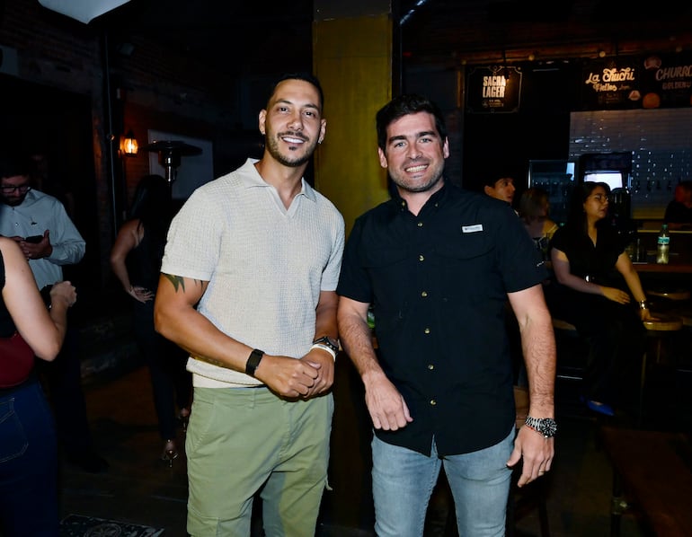 Dos hombres sonrientes, uno con camiseta clara y tattoo visible, en bar moderno con decoración de ladrillo expuesto.