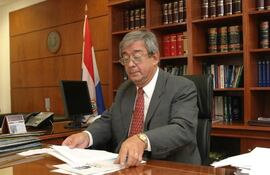 Hombre canoso con gafas en traje oscuro, sentado en escritorio con computadora y documentos, examina papeles en ambiente oficial.
