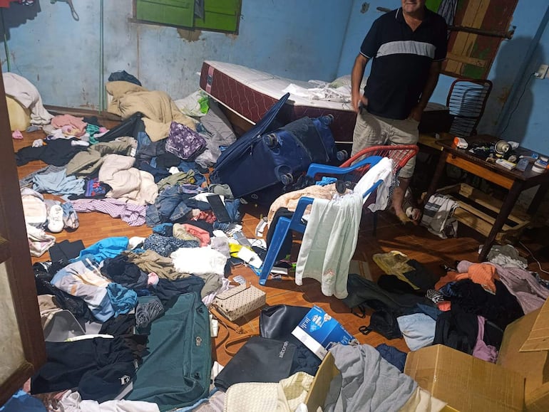 Hombre de pie en habitación desordenada, vestido con camiseta oscura y pantalones cortos, rodeado de ropa y objetos diversos.