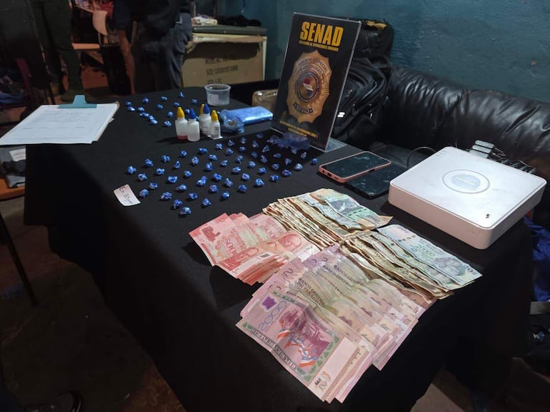Mesa con billetes y paquetes envueltos en papel azul, cartel de SENAD visible en un ambiente operativo.