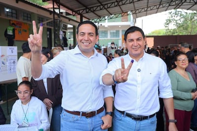 Hombres felices en camisa a rayas y blanca, muestran dedos en 'V' en ambiente festivo de votación.
