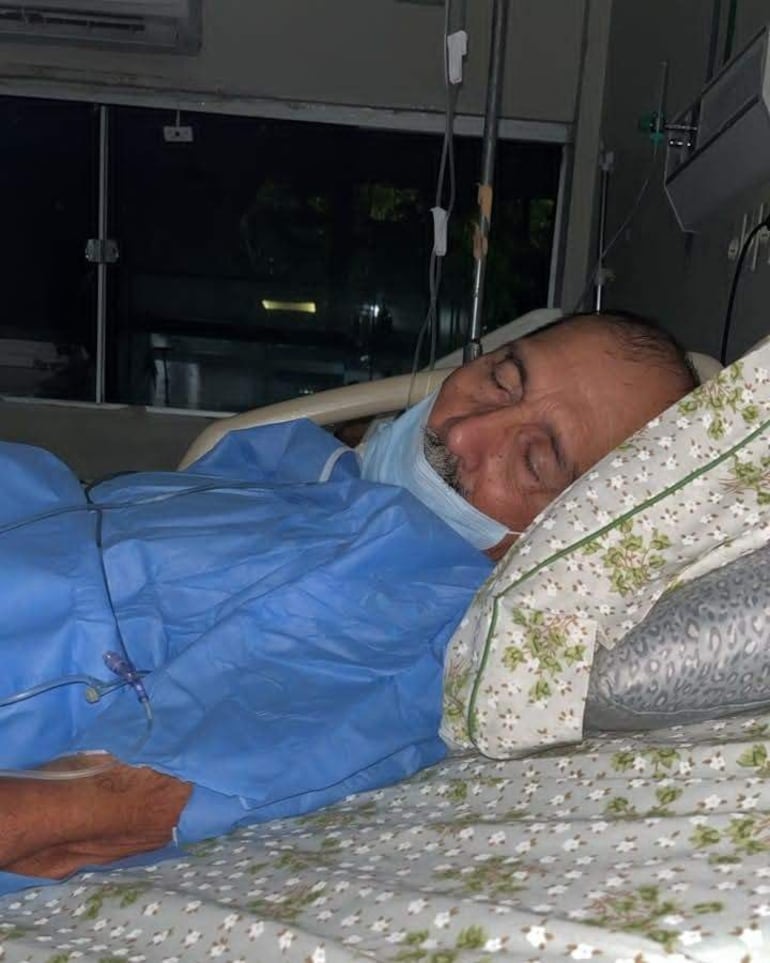 Andrés Alberto Méndez López, de 68 años, acostado en cama de hospital, cubierto con bata azul, usando mascarilla en entorno clínico.