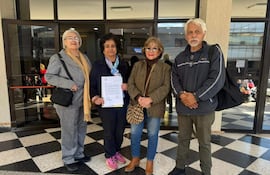 Cuatro personas, incluyendo a una mujer en el centro con un documento, posan frente a un edificio moderno con gran luz natural.