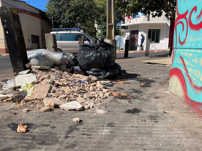 Montón de basura y escombros en la calle, con un vehículo estacionado al fondo y graffiti colorido en la pared.