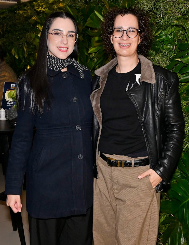 Mujeres sonrientes en un ambiente acogedor, una con abrigo oscuro y pañuelo, otra con chaqueta de cuero y pantalones claros.