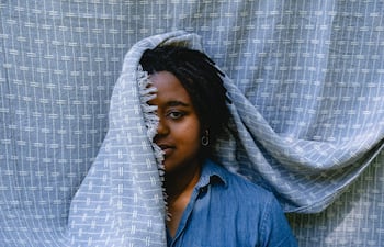 Gabriele Leite con cabello rizado, cubierto por una manta gris, viste una camisa de denim en un entorno sencillo.