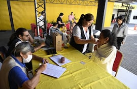 Mujer con uniforme blanco inyecta a mujer de cabello rizado sentada. Hombres con computadora y documentación en ambiente amarillo y rojo.