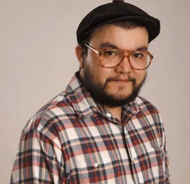 Hombre con barba, gafas grandes y sombrero tipo boina, lleva camisa de cuadros en fondo neutro.