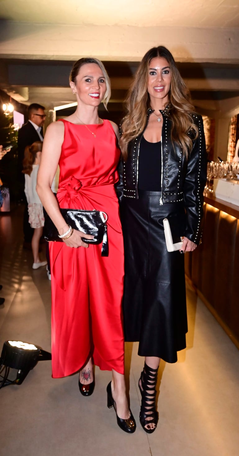 Dos mujeres, una con vestido rojo y otra en blusa negra y chaqueta de cuero, posan en una celebración con cócteles de fondo.