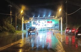 La carretera mojada y varios vehículos detenidos, con una pantalla publicitaria iluminada al fondo en una noche lluviosa.