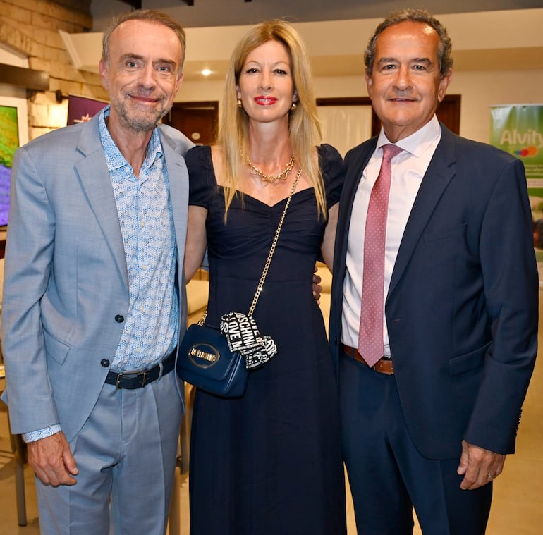 Tres personas sonrientes en un evento elegante: una mujer con cartera de diseño y dos hombres bien vestidos a su lado.