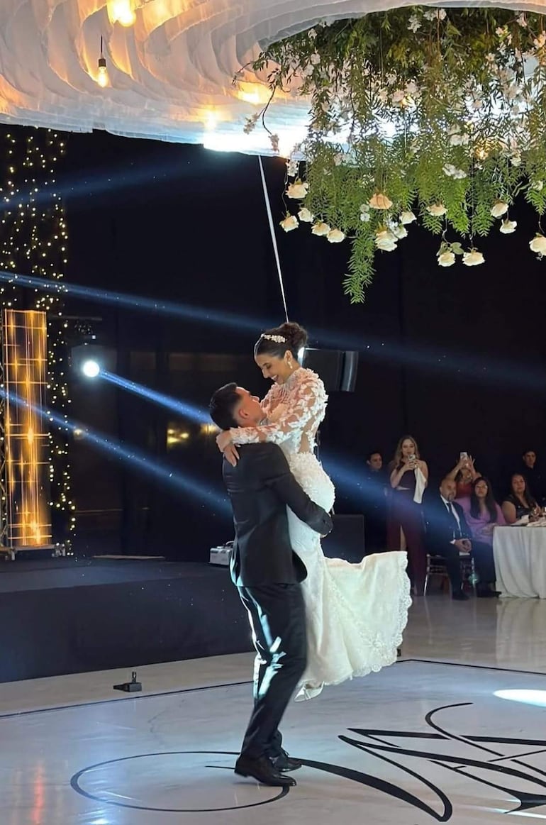Pareja sonriente bailando en su boda, él con traje oscuro y ella en vestido blanco con encaje, ambiente festivo con decoraciones florales.