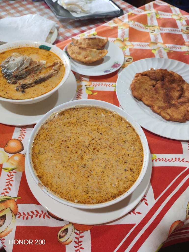 Mesa decorada con mantel rojo, tazón de caldo espeso con pescado, carne frita y empanadas doradas.