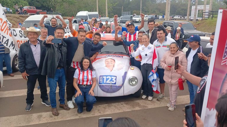 Grupo de personas con camisetas de la bandera paraguaya y brazos levantados, rodeados de un colorido auto y carteles de campaña.