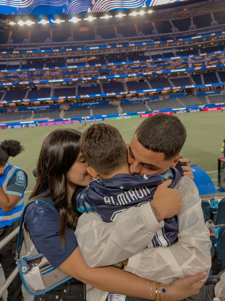 Adulto masculino y femenino abrazan a un niño con camiseta 'Almirón' en un estadio. Guardian de seguridad al fondo.