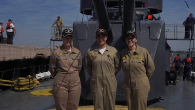 Tres mujeres uniformadas sonríen en un barco, con overoles y insignias en un entorno naval lleno de equipos.