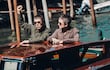 Noel Gallagher con prenda verde expresando emociones y Liam Gallagher en camisa clara, viajando en un taxi acuático por aguas tranquilas.