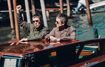 Noel Gallagher con prenda verde expresando emociones y Liam Gallagher en camisa clara, viajando en un taxi acuático por aguas tranquilas.