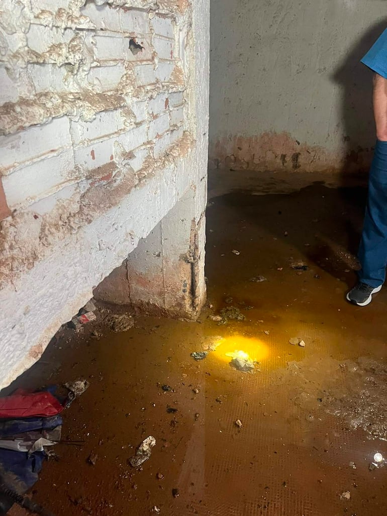 Persona con camiseta azul de pie en un sótano inundado, rodeada de agua estancada y escombros, bajo una luz tenue.