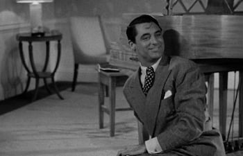 Hombre de pie, traje gris y corbata de puntos, sonriendo en entorno elegante con muebles clásicos y lámpara.
