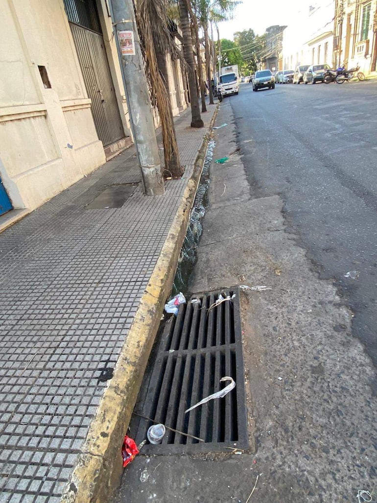 Calle desierta con vereda de baldosas, residuos en el suelo y agua estancada cerca de una alcantarilla.
