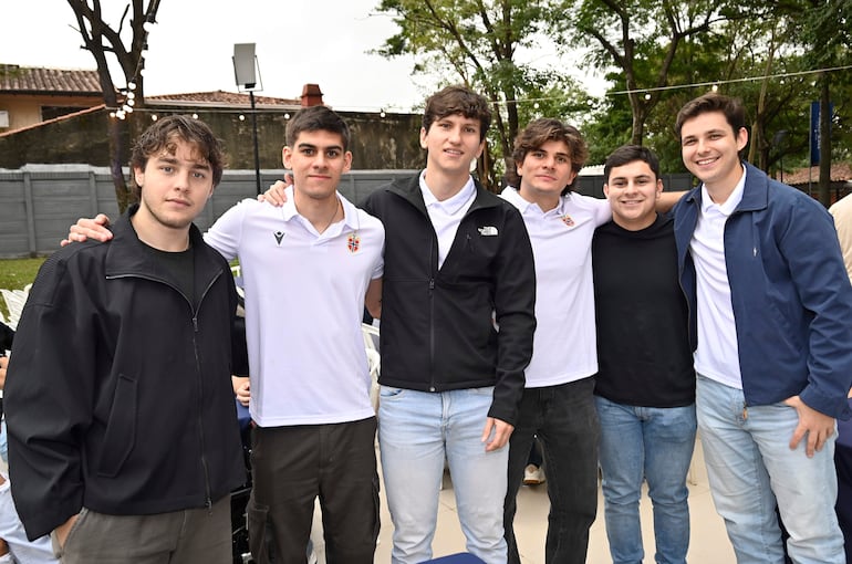 Seis jóvenes sonriendo, de pie en un ambiente exterior con árboles, algunos vistiendo camisetas blancas, todos con posturas relajadas.