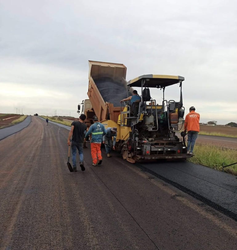 Seis trabajadores en un entorno rural, manipulando materiales cerca de una máquina de asfalto bajo un cielo nublado.