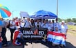 Grupo de aproximadamente 20 personas con camisetas blancas y pancarta de 'SINA MED', sosteniendo la bandera paraguaya y sonriendo.