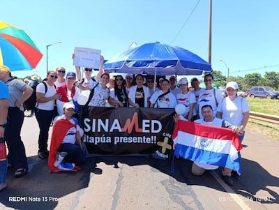 Grupo de aproximadamente 20 personas con camisetas blancas y pancarta de 'SINA MED', sosteniendo la bandera paraguaya y sonriendo.