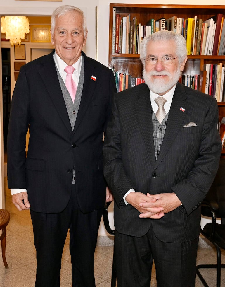 Dos hombres de pie en un ambiente iluminado con estanterías de libros, uno en traje oscuro con corbata rosa y el otro en traje negro con corbata plateada.