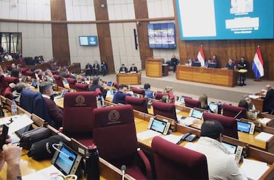 Personas sentadas en mesas, usando dispositivos electrónicos, ambiente formal con banderas de Paraguay al fondo.