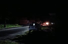 Calle oscurecida por la falta de luz, con un automóvil blanco estacionado bajo la tenue iluminación de vehículos lejanos.