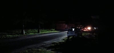 Calle oscurecida por la falta de luz, con un automóvil blanco estacionado bajo la tenue iluminación de vehículos lejanos.