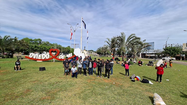 Indígenas en parque durante manifestación, algunos sentados y otros de pie, con banderas y un cartel que dice 'OVIEDO DE MI'.