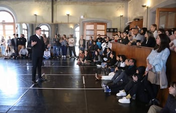 Hombre gesticulando en traje oscuro, mientras público atento escucha en un ambiente iluminado con detalles clásicos.