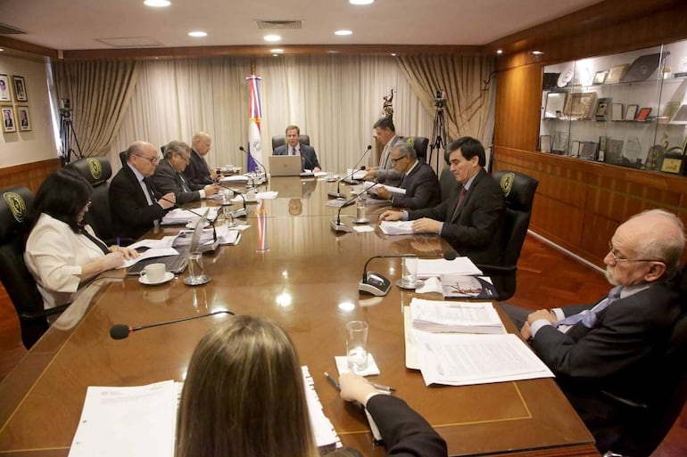 Ocho personas sentadas en mesa de reunión, con documentos y micrófonos. Ambiente formal con bandera del Paraguay y trofeos.