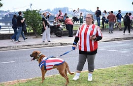 Mujer de cabello largo con camiseta a rayas del Club Olimpia pasea un perro, en medio de una multitud en la costanera.