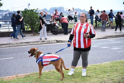 Mujer de cabello largo con camiseta a rayas del Club Olimpia pasea un perro, en medio de una multitud en la costanera.
